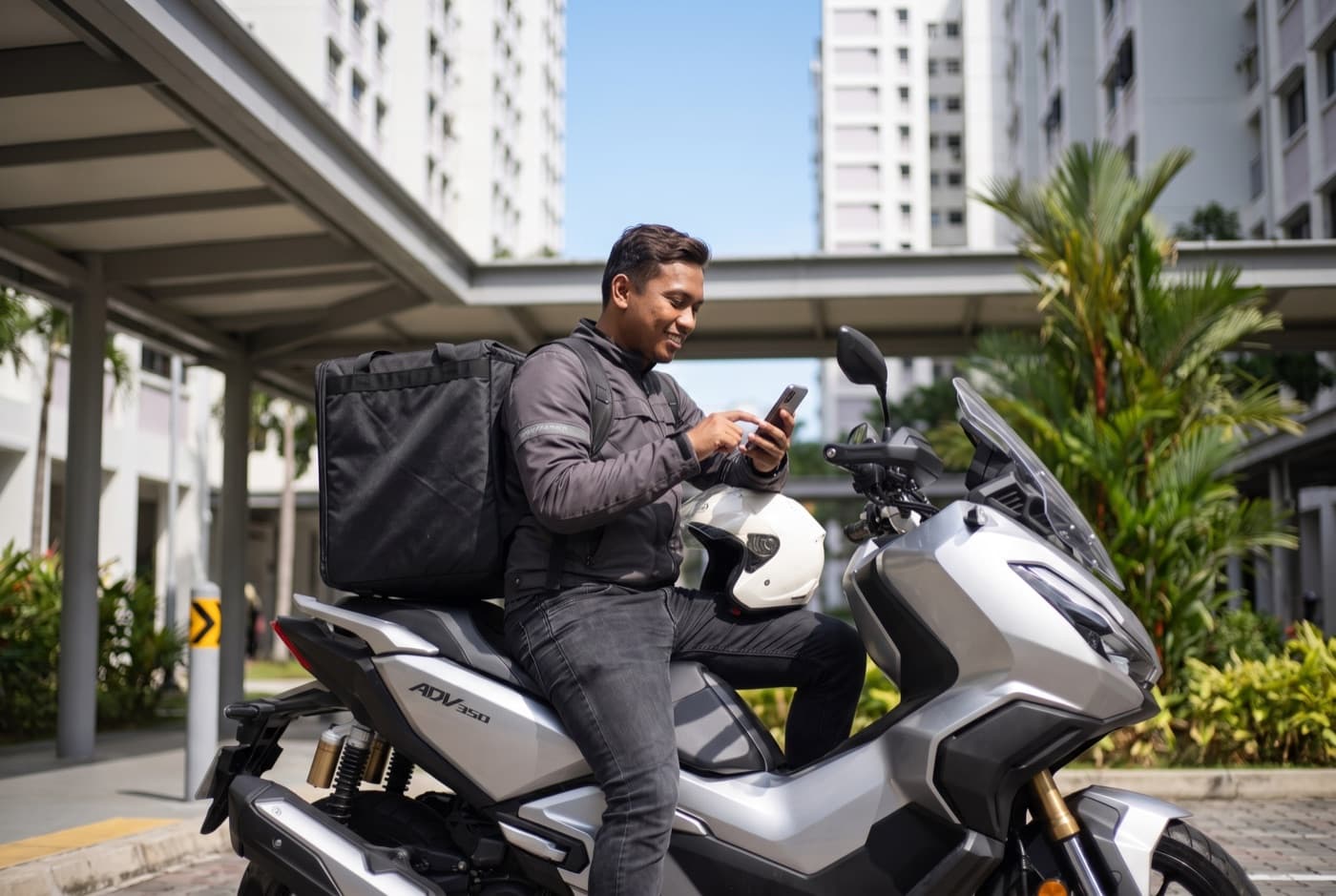 A rider tapping his phone to confirm a booking, seated on his bike under an HDB covered walkway